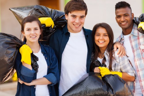 A group of four young adults standing outdoors on a paved surface, holding large black rubbish bags filled with waste. They are smiling and looking at the camera, with some of them wearing yellow gloves. The background is slightly blurred but suggests a suburban or urban environment with buildings and greenery. The individuals are dressed casually, and the scene depicts an activity related to rubbish collection or waste disposal, aligning with professional rubbish removal services such as those offered by Office Clearance Tufnell Park. The overall atmosphere is positive and shows teamwork in waste management efforts, suitable for a page about office clearance and rubbish removal services in the Tufnell Park area, London postcodes.