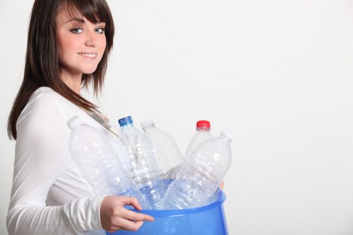 A young woman with shoulder-length brown hair and a light complexion is smiling slightly while holding a blue recycling bin filled with clear plastic bottles, some with red or blue caps, in an indoor setting with a plain white background. The bottles vary in size and shape, and appear empty. The woman is dressed in a white long-sleeved top, and her posture suggests she is about to dispose of or recycle the bottles. This image relates to rubbish and waste management services, highlighting the collection and recycling of plastic waste, which is a common task for companies like Office Clearance Tufnell Park. The clear, well-lit environment emphasizes cleanliness and disposal activities, supporting local waste removal and recycling efforts in the Tufnell Park area.