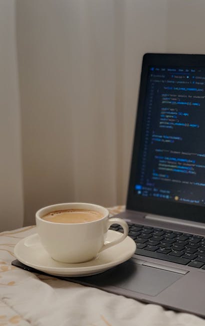 A white ceramic cup filled with coffee or tea sits on a matching white saucer, placed on a light-colored surface such as a bed or table. Adjacent to the cup, slightly to the right, is an open silver l