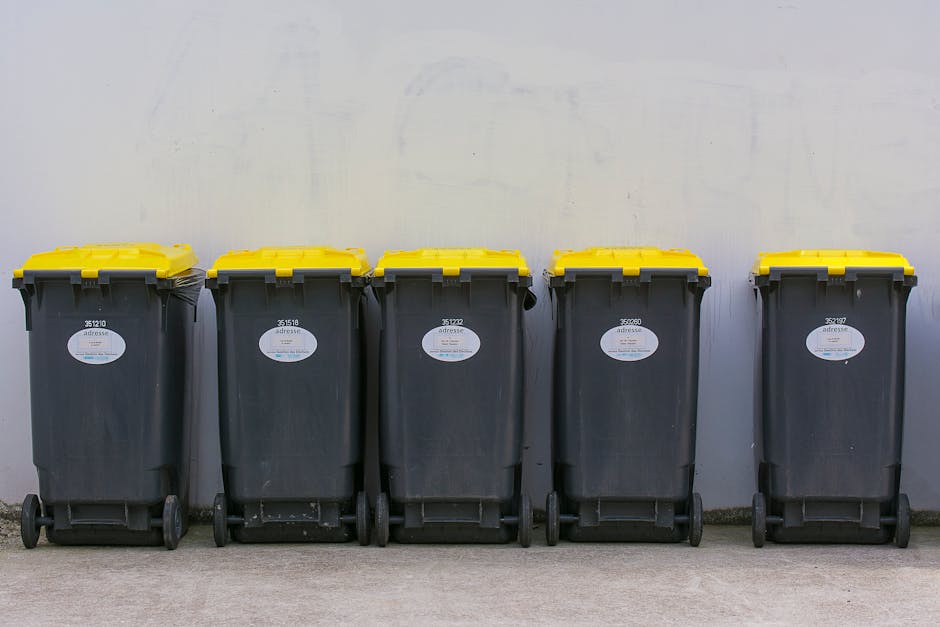 A middle-aged woman with short dark hair, dressed in a black T-shirt, is using a wooden-handled grabber tool to dispose of waste into a cylindrical stainless steel litter bin on a paved sidewalk. She