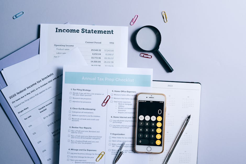 A flat lay of a workspace featuring financial documents related to tax preparation, including an 'Income Statement' and an 'Annual Tax Prep Checklist', spread out on a light grey surface. The document